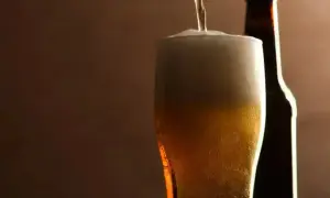 Birra pils che si versa in un bicchiere, con una bottiglia sullo sfondo. Schiuma abbondante e colore dorato invitante.