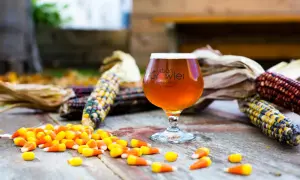 Bicchiere di birra ambrata con logo Urban Growler, circondato da pannocchie colorate e caramelle a forma di mais. Un omaggio autunnale alla birra al mais.