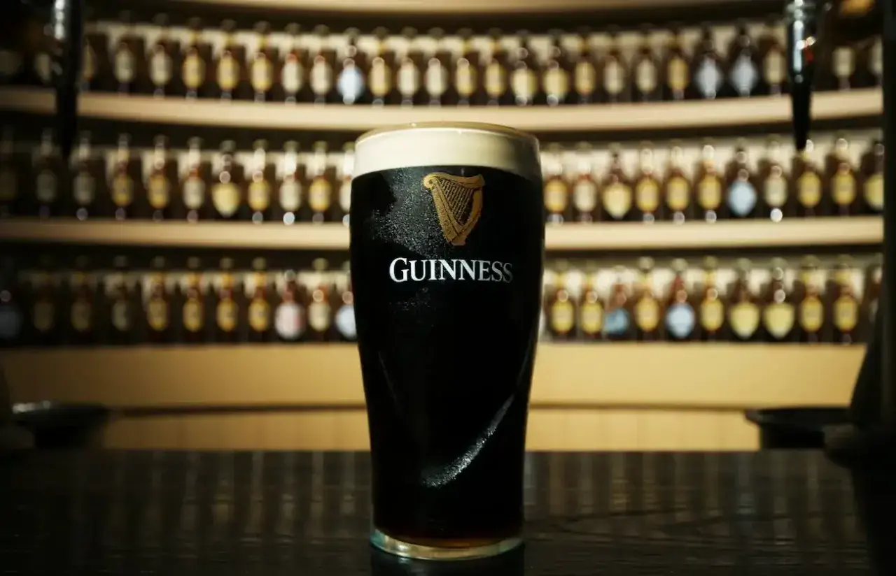 Un bicchiere di Guinness, con il suo logo iconico, pronto per essere gustato. Dietro, una parete di bottiglie, quasi una famiglia di Guinness in attesa.