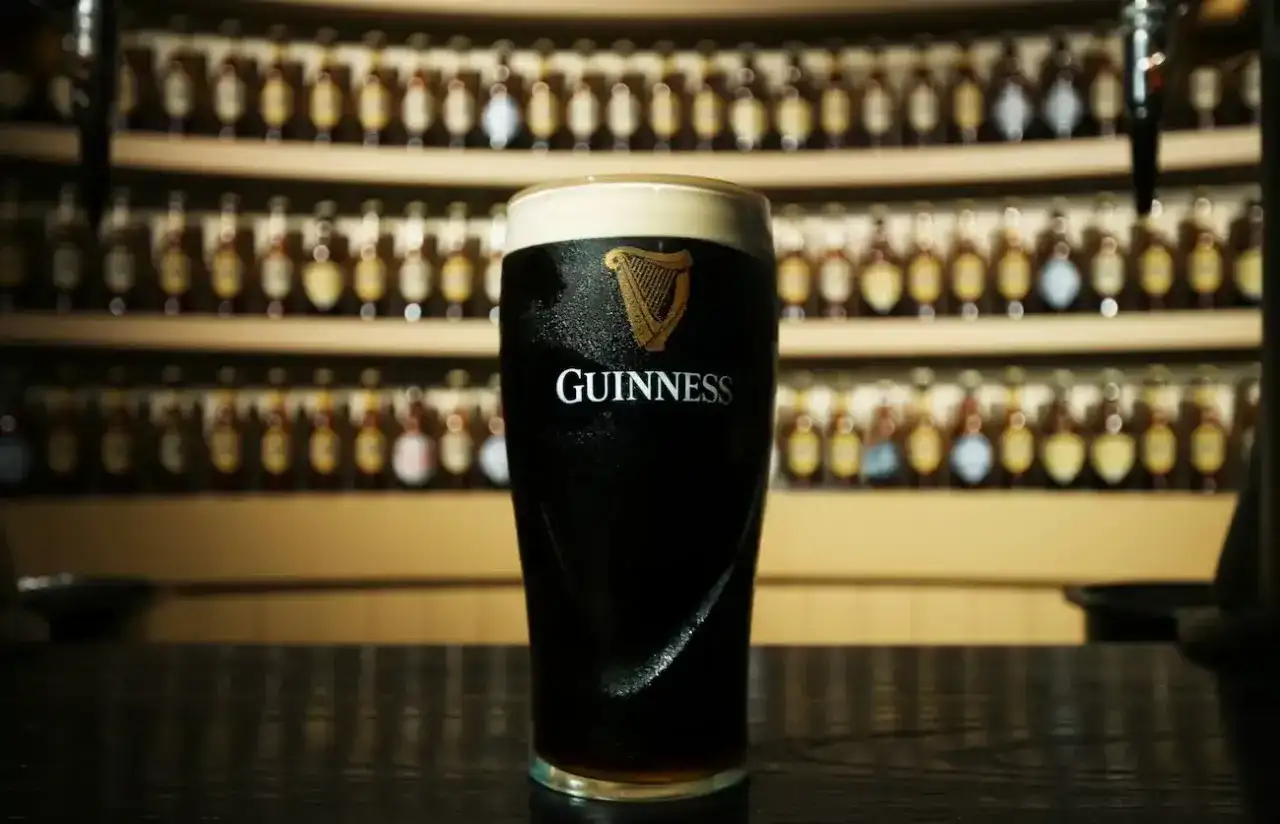 Un bicchiere di Guinness, con il suo logo iconico, pronto per essere gustato. Dietro, una parete di bottiglie, quasi una famiglia di Guinness in attesa.