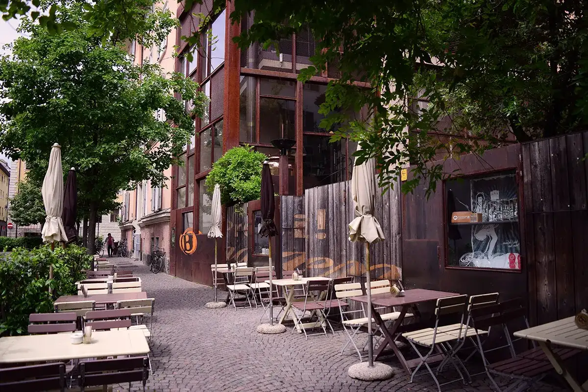 Tavoli all'aperto e ombrelloni di fronte al Batzen Bräu, con un'architettura industriale e un'atmosfera rilassata.