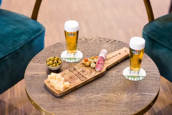 Due bicchieri di birra ale Heineken con salumi, formaggio e olive su un tavolino di legno.