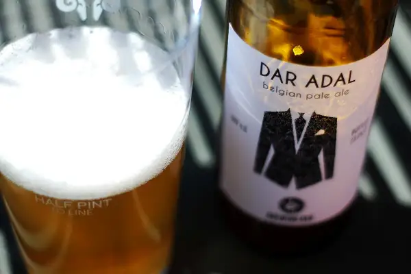 Bicchiere di birra dorata con schiuma bianca e bottiglia di Dar Adal belgian pale ale con etichetta a forma di giacca.