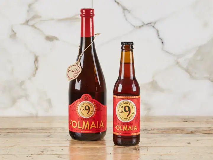 Due bottiglie di birra ambrata "La 9" del Birrificio L'Olmaia, una da 750ml e una più piccola, su un tavolo di legno.