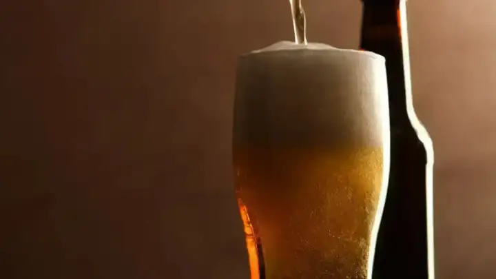 Birra pils che si versa in un bicchiere, con una bottiglia sullo sfondo. Schiuma abbondante e colore dorato invitante.